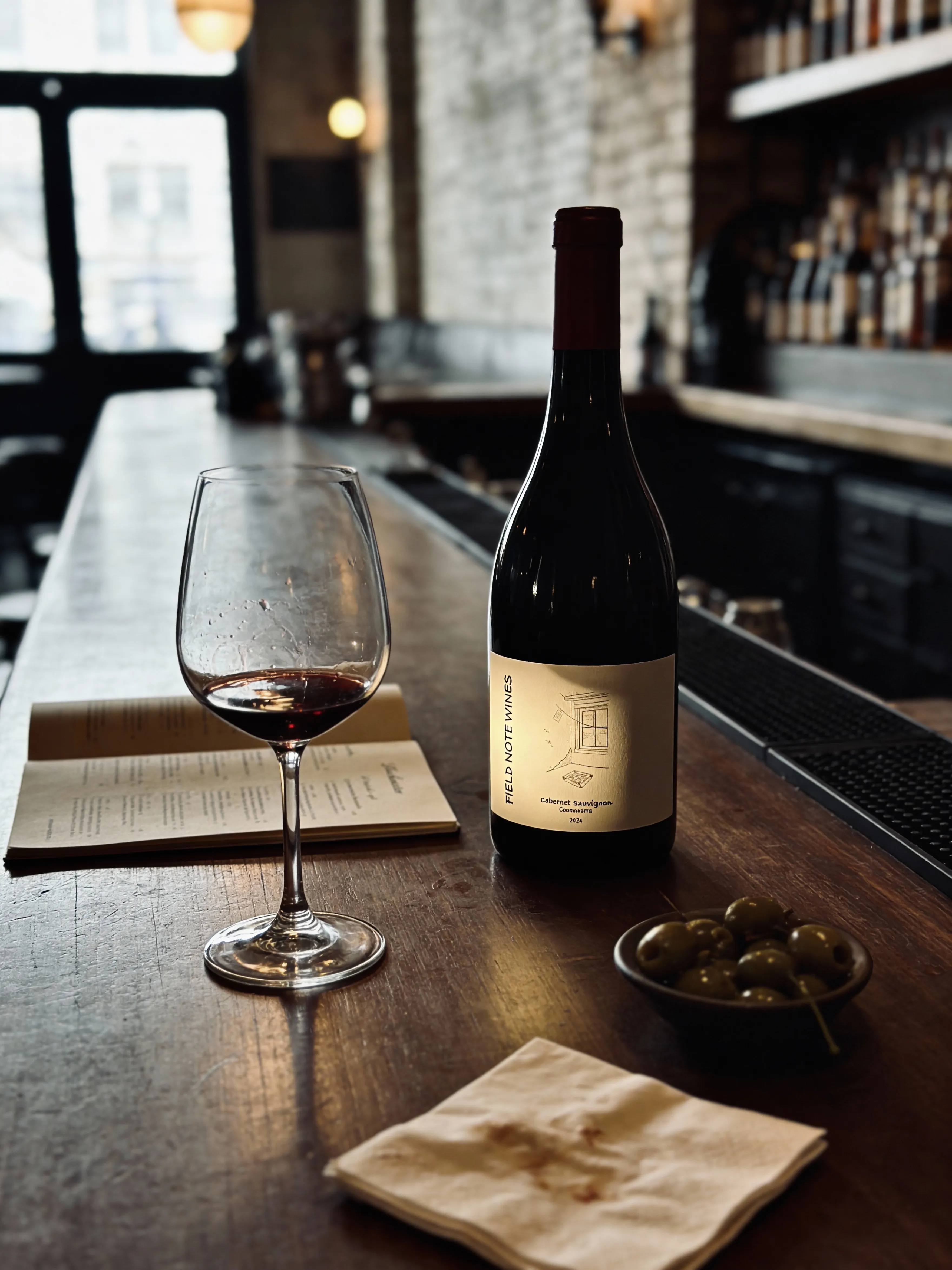 FIELD NOTE bottle and wine glass on a dark wooden bar with small table objects